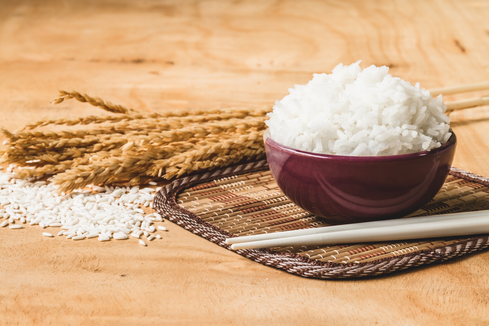 Cooked rice in bowl with raw rice grain and dry rice plant on wooden table background.